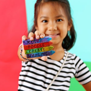May include: A young person holds a stack of colorful crayon-shaped art supplies. Each crayon is a different color and has a word on it: "COLOR", "DOODLE", "CREATE", and "IMAGINE". The background is split between red and blue.