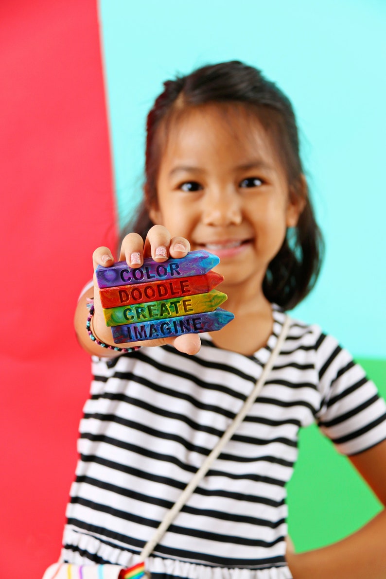 May include: A young person holds a stack of colorful crayon-shaped art supplies. Each crayon is a different color and has a word on it: "COLOR", "DOODLE", "CREATE", and "IMAGINE". The background is split between red and blue.