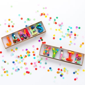 May include: Two sets of colorful crayon letters spelling out the names "JAMES" and "KYLIE" in a gold box on a white background with confetti.