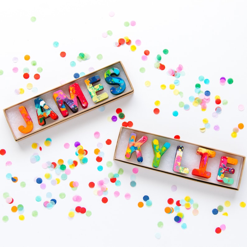 May include: Two sets of colorful crayon letters spelling out the names "JAMES" and "KYLIE" in a gold box on a white background with confetti.