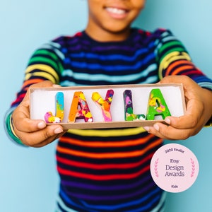 May include: A child holds a box with the name "JAYLA" spelled out in colorful crayon letters. The box is made of cardboard and has a white interior. The child is wearing a rainbow striped shirt. The image also includes a pink circle with the text "2020 Finalist Etsy Design Awards Kids".