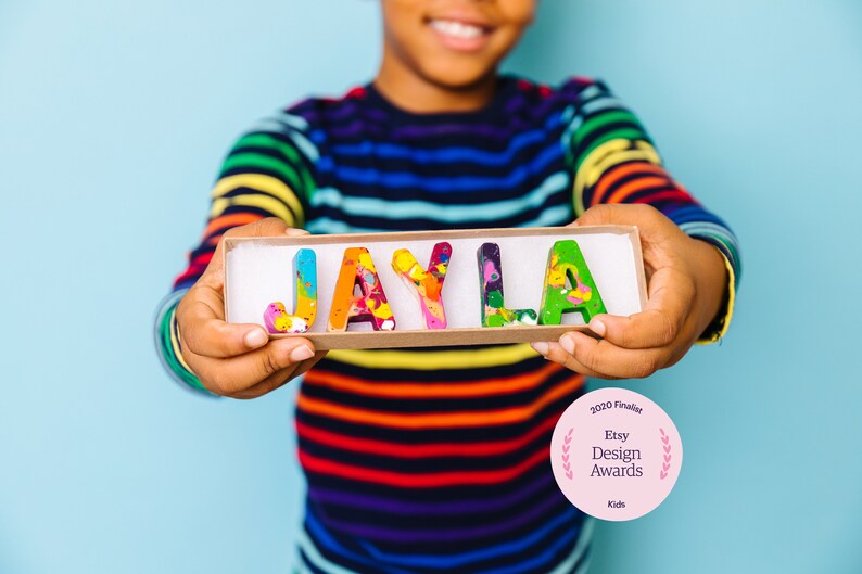 May include: A child holds a box with the name "JAYLA" spelled out in colorful crayon letters. The box is made of cardboard and has a white interior. The child is wearing a rainbow striped shirt. The image also includes a pink circle with the text "2020 Finalist Etsy Design Awards Kids".