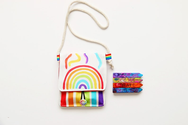 May include: A white crossbody bag with a rainbow design and a matching rainbow strap. The bag has a colorful striped bottom and a lollipop button closure. Next to the bag are four colorful crayons with the words "COLOR DOODLE CREATE IMAGINE".