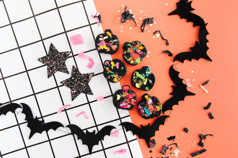 Set of 4 Halloween crayons in the shape of a pumpkin. The base color is black with brighter and neon flecks of color on top. Crayon set comes in a clear top, Kraft colored box with a band around the outside.