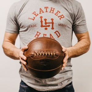 May include: A person holding a brown leather basketball with white stitching and the text "Leather Head" embossed on the side.