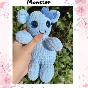 Op de afbeelding: Lichtblauw gehaakt knuffel met grote zwarte ogen en een kleine glimlach. De knuffel heeft een strik op het hoofd en is gelabeld "Olivia the Flower Monster". De afbeelding bevat ook de tekst "Crochet pattern".