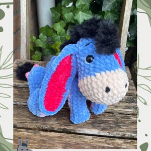 Op de afbeelding: Een blauw gehaakt ezelknuffel met roze binnenoren en een zwarte manen en staart. De tekst "Dustin the Donkey" staat boven de ezel. De tekst "Crochet pattern by @joolscrochetplushies" staat eronder.