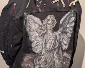 Puede incluir: Chaqueta vaquera negra con un diseño de ángel pintado a mano en la espalda. La chaqueta tiene detalles de cinta blanca y ojales rosas. El ángel está representado en tonos grises y blancos, con alas detalladas.