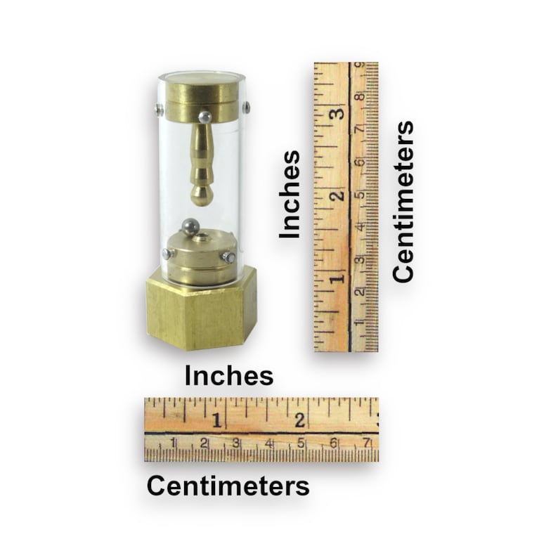 May include: A brass and glass perpetual motion device with a small ball rolling inside a clear cylinder. The device is approximately 5 cm tall and is sitting on a wooden surface.