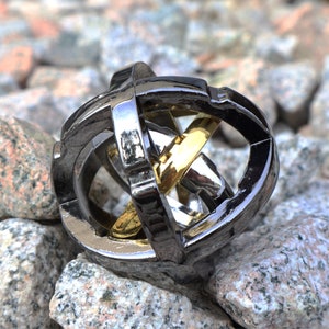 May include: A silver metal puzzle with a gold ring inside. The puzzle is made of interlocking rings and is designed to be challenging to solve.
