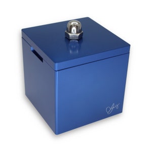 Peut inclure: Une boîte à énigmes en bois bleu avec un bouton argenté sur le dessus. La boîte porte une petite signature gravée sur le devant.