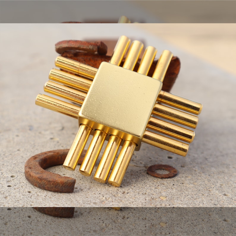 May include: A gold metal puzzle with a square center and eight rods extending out from the center. The puzzle is lying on a light brown surface.