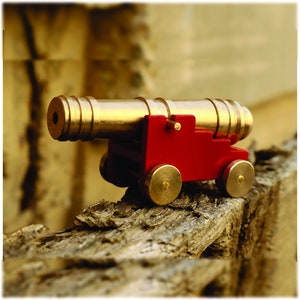 May include: A small, brass cannon on a red wooden carriage. The cannon is mounted on wheels and has a barrel with a flared muzzle. The cannon is sitting on a piece of weathered wood.