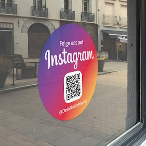 Op de afbeelding: Een ronde sticker met een verloop van paars, rood en oranje. De sticker heeft het woord "Instagram" in het wit, een QR-code en de tekst "@DeinNutzername". De sticker zit op een glazen raam.
