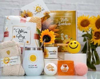 May include: A gift basket filled with items themed around sunshine and warmth. It includes a white mug with a yellow sun and the text "A Cup of Sunshine", a white candle with the text "Sending You Sunshine", a white bath bomb, a yellow bath bomb, a yellow smiley face, a white towel with the text "Sending Sunshine and Hugs", a white blanket with the text "A Warm Hug", and a yellow and white box with the text "Hello Sunshine".