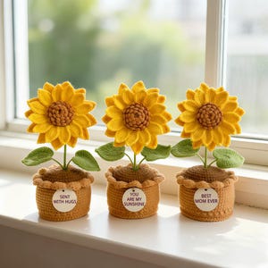 May include: Three handmade crochet sunflowers in small brown pots sit on a windowsill. The yellow sunflowers have brown centers and green leaves. Each pot has a small tag with text: "SENT WITH HUGS", "YOU ARE MY SUNSHINE", and "BEST MOM EVER".