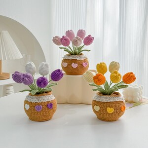 May include: Three crocheted flower arrangements in pots. Each pot is decorated with a white lace trim and small heart accents. The flowers are pink, purple, and yellow.
