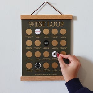 May include: A West Loop restaurant scratch-off poster with a dark green background and gold lettering. The poster is framed in light wood and includes a list of restaurants with scratch-off circles. A hand is scratching off a circle.