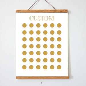 May include: A white print with the word "CUSTOM" in gold lettering, featuring rows of gold circles. The print is displayed in a wooden frame, hanging against a white wall.