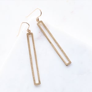 May include: A pair of gold-colored, rectangular drop earrings. Each earring features a long, open rectangle design with a hook for pierced ears. The earrings are displayed on a white marble surface.