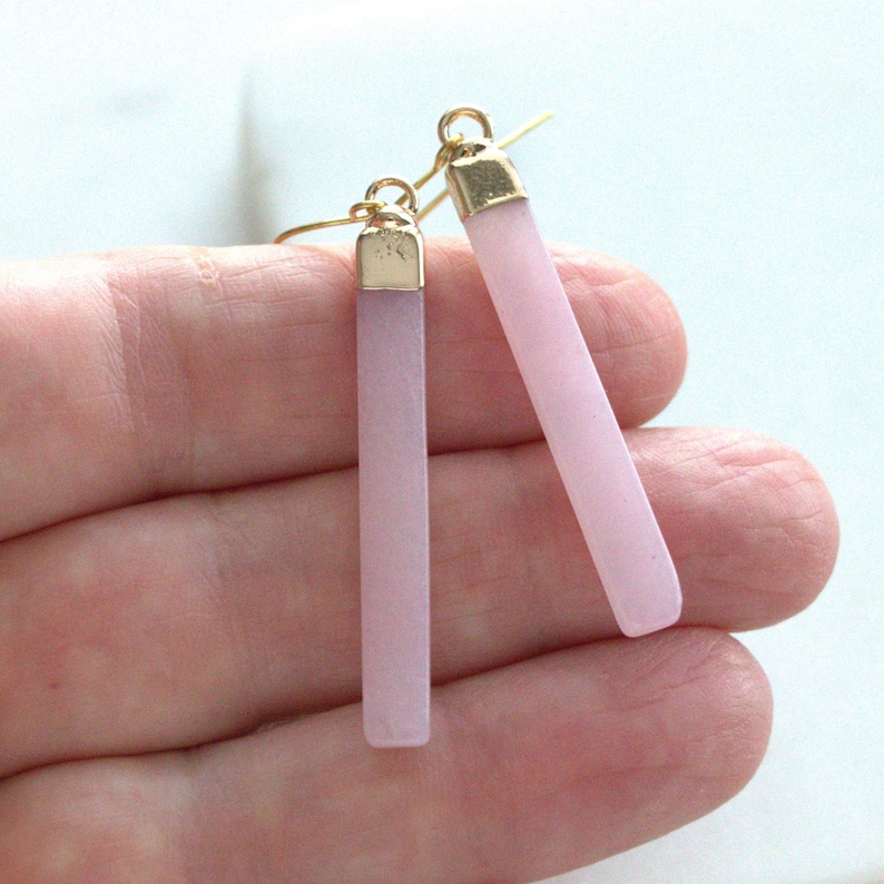 May include: A pair of light pink bar earrings with gold-colored caps and ear wires. The earrings are rectangular in shape and have a matte finish. The earrings are shown against a light background.