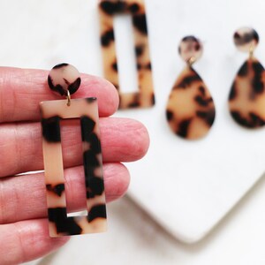 May include: A pair of brown and black tortoise shell earrings. One earring is a rectangle with a rectangular cutout in the center. The other earring is a teardrop shape.