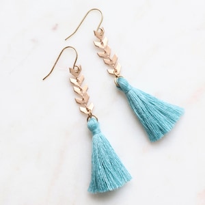 May include: A pair of gold-colored earrings with a chevron design and light blue tassels. The earrings have a hook closure and are displayed on a white surface. The tassels are a soft, light blue color.