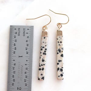May include: A pair of dangle earrings with a rectangular, speckled design. The earrings are approximately 2 inches long, with a gold-colored hook and a gold-colored cap at the top of the stone. The stone has a cream background with black spots.
