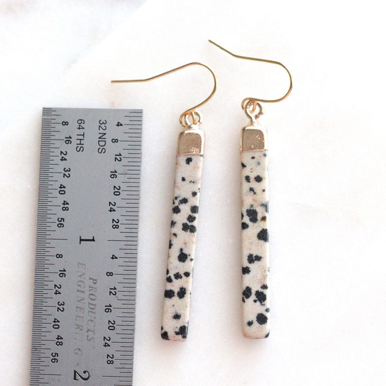 May include: A pair of dangle earrings with a rectangular, speckled design. The earrings are approximately 2 inches long, with a gold-colored hook and a gold-colored cap at the top of the stone. The stone has a cream background with black spots.