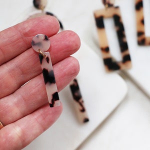 May include: A pair of dangle earrings with a tortoiseshell pattern. The earrings are made of a white and black material and have a gold-colored metal hook.
