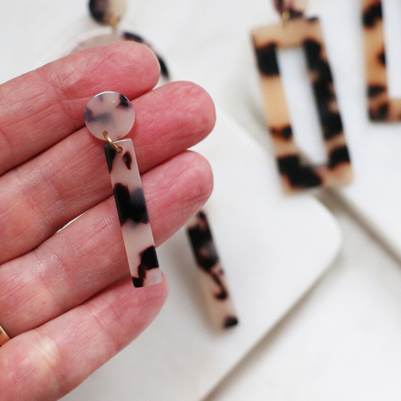 May include: A pair of dangle earrings with a tortoiseshell pattern. The earrings are made of a white and black material and have a gold-colored metal hook.