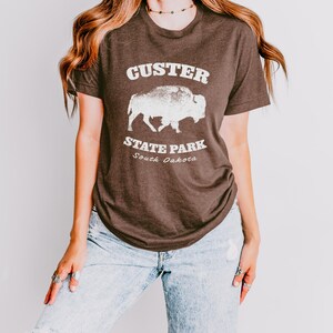 Model is wearing a Heather brown t-shirt with the text "Custer State Park" in white and a bison graphic. The shirt is paired with jeans for a casual look. This athletic style tee is made of soft and cozy cotton and features the Bella and Canvas flattering fit and durability. These shirts are short sleeve crew neck style and make great adventure souvenirs for a hiking or camping trip to South Dakota as well as birthday, anniversary, Christmas, Mothers and Fathers Day gifts.