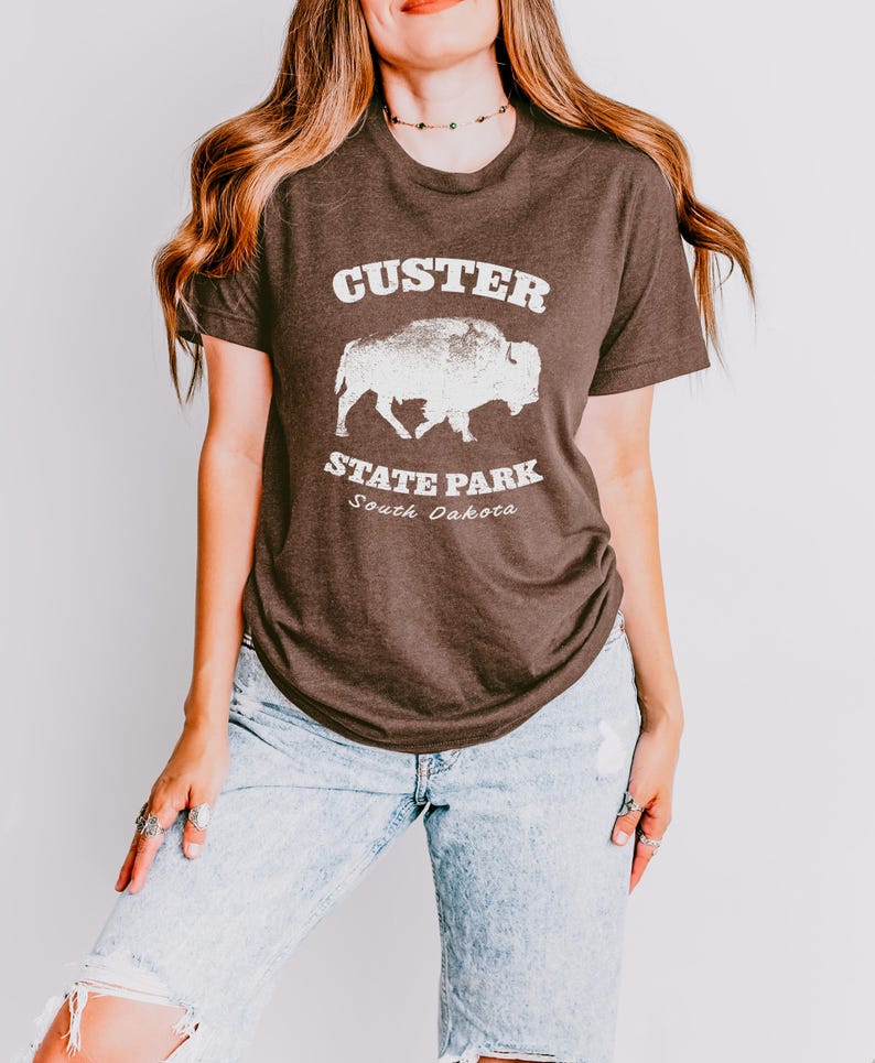 Model is wearing a Heather brown t-shirt with the text "Custer State Park" in white and a bison graphic. The shirt is paired with jeans for a casual look. This athletic style tee is made of soft and cozy cotton and features the Bella and Canvas flattering fit and durability. These shirts are short sleeve crew neck style and make great adventure souvenirs for a hiking or camping trip to South Dakota as well as birthday, anniversary, Christmas, Mothers and Fathers Day gifts.
