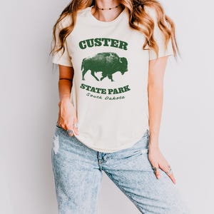 Model is wearing an off white t-shirt with the text "Custer State Park" in dark green and a bison graphic. The shirt is paired with jeans for a casual look. This athletic style tee is made of soft and cozy cotton and features the Bella and Canvas flattering fit and durability. These shirts are short sleeve crew neck style and make great adventure souvenirs for a hiking or camping trip to South Dakota as well as birthday, anniversary, Christmas, Mothers and Fathers Day gifts.