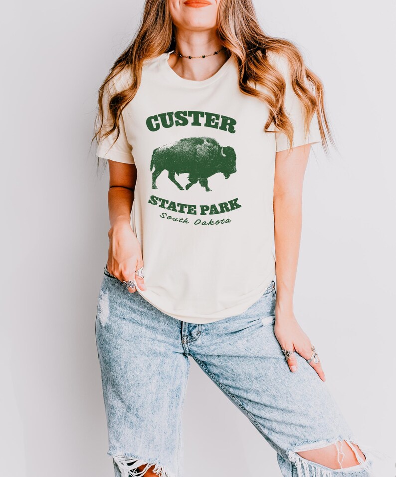 Model is wearing an off white t-shirt with the text "Custer State Park" in dark green and a bison graphic. The shirt is paired with jeans for a casual look. This athletic style tee is made of soft and cozy cotton and features the Bella and Canvas flattering fit and durability. These shirts are short sleeve crew neck style and make great adventure souvenirs for a hiking or camping trip to South Dakota as well as birthday, anniversary, Christmas, Mothers and Fathers Day gifts.