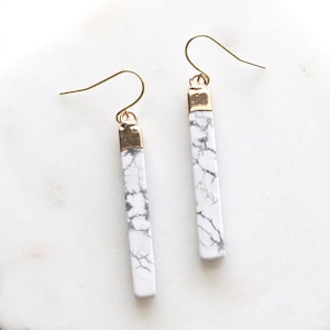 May include: A pair of dangle earrings featuring rectangular white marble stones with gray veining. The earrings have gold-colored metal accents and ear wires. These earrings are a stylish accessory.