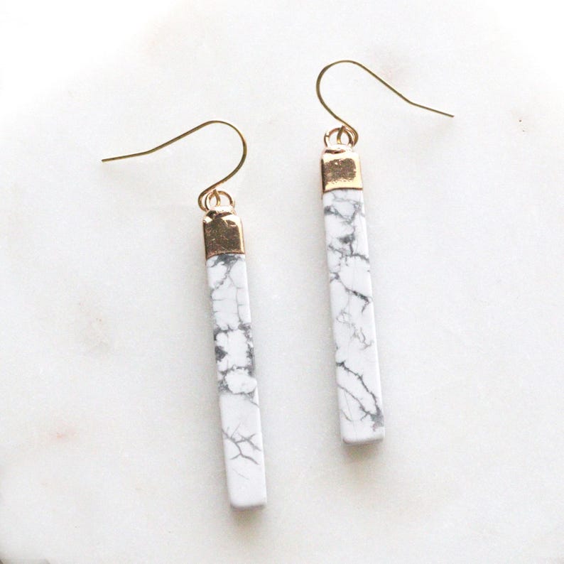 May include: A pair of dangle earrings featuring rectangular white marble stones with gray veining. The earrings have gold-colored metal accents and ear wires. These earrings are a stylish accessory.