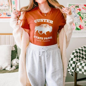 Model is wearing an autumn orange t-shirt with the text "Custer State Park" in white and a bison graphic. The shirt is paired with sweatpants for a casual look. This athletic style tee is made of soft and cozy cotton and features the Bella and Canvas flattering fit and durability. These shirts are short sleeve crew neck style and make great adventure souvenirs for a hiking or camping trip to South Dakota as well as birthday, anniversary, Christmas, Mothers and Fathers Day gifts.