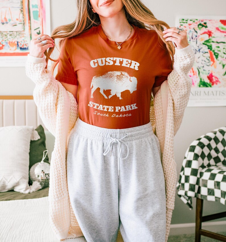 Model is wearing an autumn orange t-shirt with the text "Custer State Park" in white and a bison graphic. The shirt is paired with sweatpants for a casual look. This athletic style tee is made of soft and cozy cotton and features the Bella and Canvas flattering fit and durability. These shirts are short sleeve crew neck style and make great adventure souvenirs for a hiking or camping trip to South Dakota as well as birthday, anniversary, Christmas, Mothers and Fathers Day gifts.