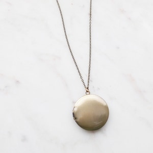 May include: A bronze-colored locket necklace with a delicate chain. The round locket is closed and has a smooth, reflective surface. The chain and locket are set against a white marble background, creating a classic and elegant look.