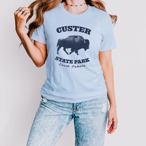 Model is wearing a light blue t-shirt with the text "Custer State Park" in dark blue and a bison graphic. The shirt is paired with jeans for a casual look. This athletic style tee is made of soft and cozy cotton and features the Bella and Canvas flattering fit and durability. These shirts are short sleeve crew neck style and make great adventure souvenirs for a hiking or camping trip to South Dakota as well as birthday, anniversary, Christmas, Mothers and Fathers Day gifts.