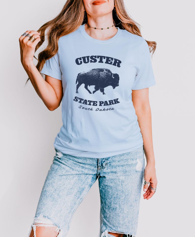 Model is wearing a light blue t-shirt with the text "Custer State Park" in dark blue and a bison graphic. The shirt is paired with jeans for a casual look. This athletic style tee is made of soft and cozy cotton and features the Bella and Canvas flattering fit and durability. These shirts are short sleeve crew neck style and make great adventure souvenirs for a hiking or camping trip to South Dakota as well as birthday, anniversary, Christmas, Mothers and Fathers Day gifts.