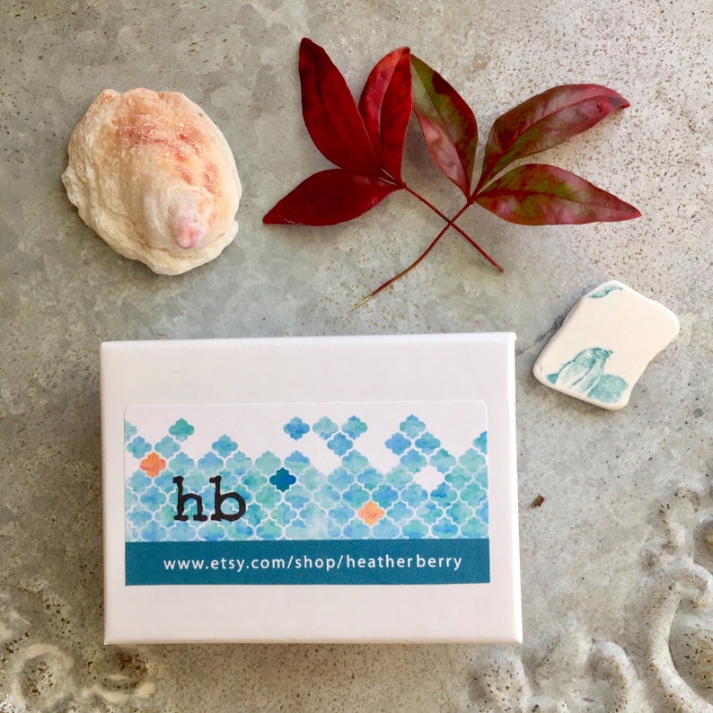 May include: A white box with a blue and orange geometric pattern and the text "hb" and "www.etsy.com/shop/heatherberry". The box is sitting on a gray surface with a seashell and two red leaves.