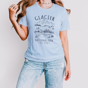 May include: A light blue t-shirt with the words "GLACIER NATIONAL PARK - EST. 1910" printed in black. The graphic depicts a mountain range, trees, and a lake. The shirt is paired with light blue jeans.