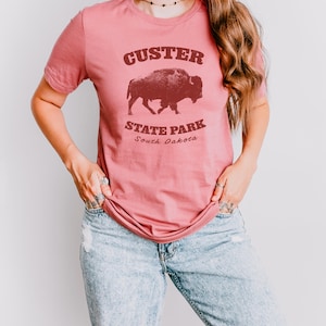 Model is wearing a medium pink t-shirt with the text "Custer State Park" in dark red and a bison graphic. The shirt is paired with jeans for a casual look. This athletic style tee is made of soft and cozy cotton and features the Bella and Canvas flattering fit and durability. These shirts are short sleeve crew neck style and make great adventure souvenirs for a hiking or camping trip to South Dakota as well as birthday, anniversary, Christmas, Mothers and Fathers Day gifts.