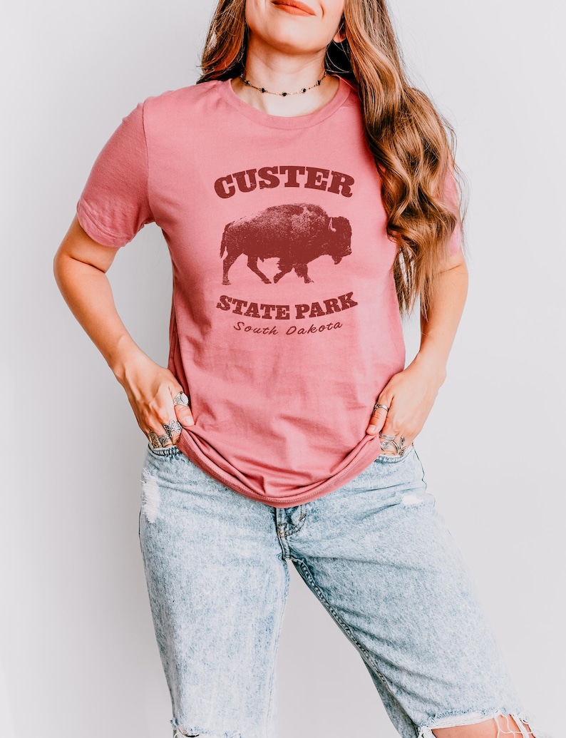 Model is wearing a medium pink t-shirt with the text "Custer State Park" in dark red and a bison graphic. The shirt is paired with jeans for a casual look. This athletic style tee is made of soft and cozy cotton and features the Bella and Canvas flattering fit and durability. These shirts are short sleeve crew neck style and make great adventure souvenirs for a hiking or camping trip to South Dakota as well as birthday, anniversary, Christmas, Mothers and Fathers Day gifts.