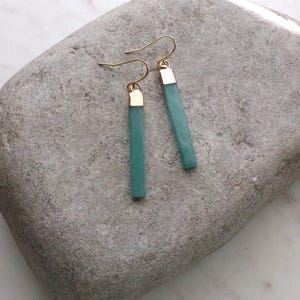May include: A pair of teal-colored, rectangular earrings with gold-colored caps and ear wires. The earrings are displayed on a gray, textured stone surface. The earrings are a simple, elegant design.