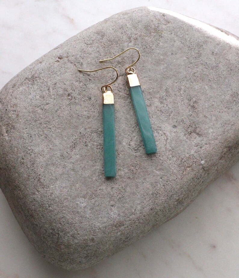 May include: A pair of teal-colored, rectangular earrings with gold-colored caps and ear wires. The earrings are displayed on a gray, textured stone surface. The earrings are a simple, elegant design.