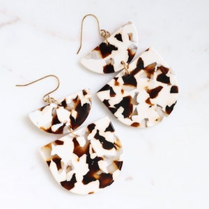 May include: A pair of white and brown tortoise shell earrings with a gold hook. The earrings are made of two semi-circles stacked on top of each other.