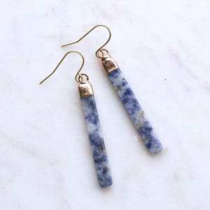 May include: A pair of dangle earrings featuring elongated, rectangular blue and white stone beads. The earrings have gold-colored metal accents at the top and gold-colored ear wires. The earrings are displayed on a white marble surface.